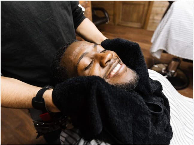 Smiling Black man relaxes as a barber wraps a warm black towel around his face.