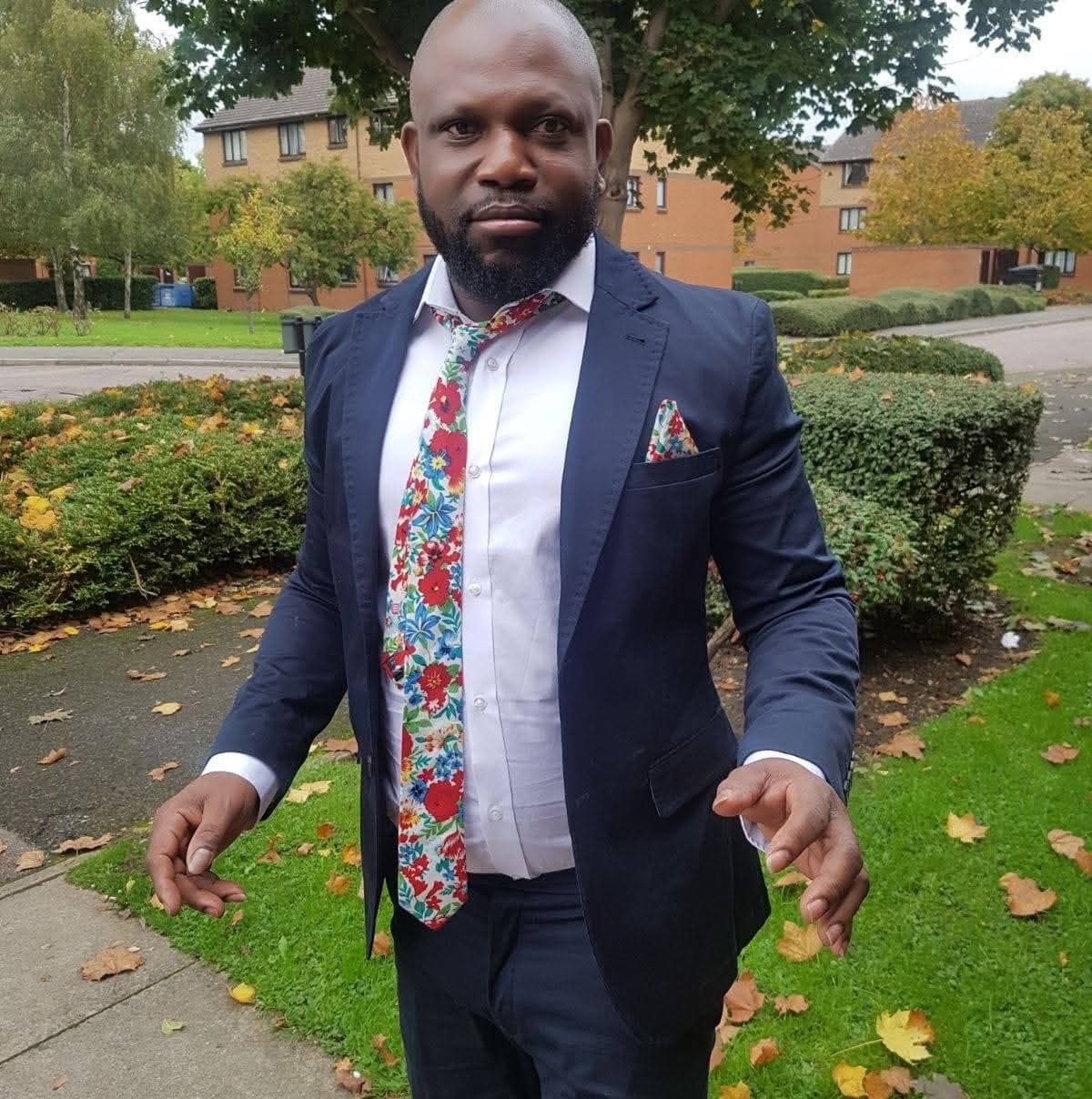Bald Black man in a navy suit and colorful floral tie standing outdoors near bushes.