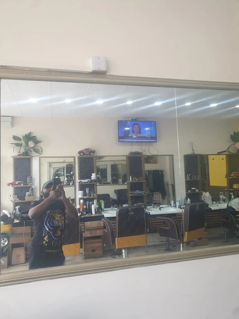 Man taking a mirror selfie in a barbershop with leather chairs and a wall-mounted TV.